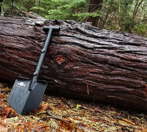 Agency 6 aluminum mini shovel black offroad overland shovel leaning against a tree trunk. This shovel bolts onto a roof rack for your 4x4 or offroad rig